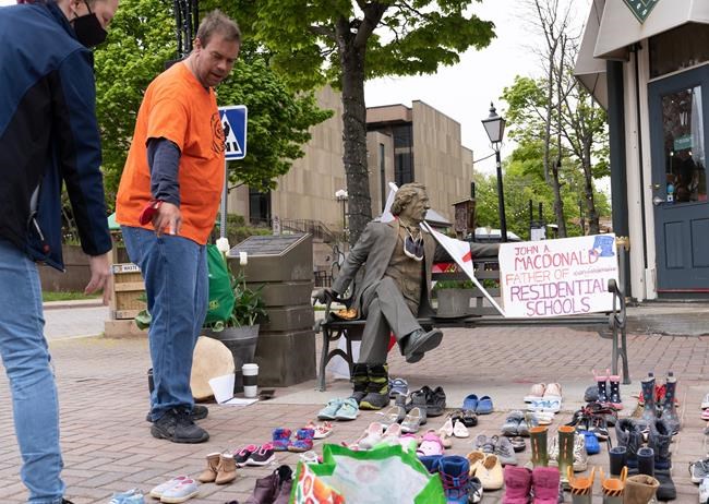 Chris Clay (right) and Leona MacDonald (left) point to some of the 215 sets of children sneakers in front of the Sir John A. MacDonald statue after 215 bodies were found at a former residential school for Indigenous in British Columbia. The sneakers were laid out on Queen Street in Charlottetown, on Monday, May 31, 2021. THE CANADIAN PRESS/John Morris
