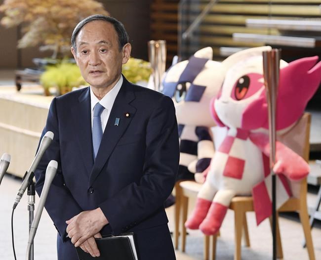 Japan's Prime Minister Yoshihide Suga speaks to media in front of the Olympics mascots at the prime minister's office in Tokyo, Thursday, July 1, 2021. Suga was still uncertain whether fans can be allowed at the Tokyo Olympics because of growing concern about rising COVID-19 cases on Thursday only three weeks before the games.(Kyodo News via AP)