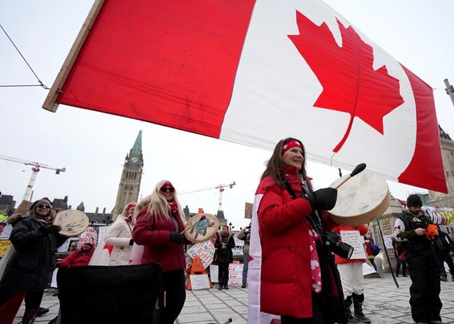 Women drum as they pass under a giant Canadian flag in front of Parliament Hill on the 20th day of a protest against COVID-19 measures that has grown into a broader antigovernment protest, in Ottawa, on Wednesday, Feb. 16, 2022. THE CANADIAN PRESS/Justin Tang