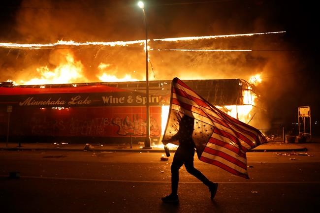 A protester carries the carries a U.S. flag upside, a sign of distress, next to a burning building Thursday, May 28, 2020, in Minneapolis. Protests over the death of George Floyd, a black man who died in police custody Monday, broke out in Minneapolis for a third straight night. (AP Photo/Julio Cortez)