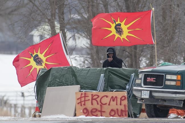 A protester stands between Mohawk Warrior Society flags at a rail blockade on the tenth day of demonstration in Tyendinaga, near Belleville, Ont., Sunday, Feb. 16, 2020. The protest is in solidarity with the Wet'suwet'en hereditary chiefs opposed to the LNG pipeline in northern British Columbia. THE CANADIAN PRESS/Lars Hagberg