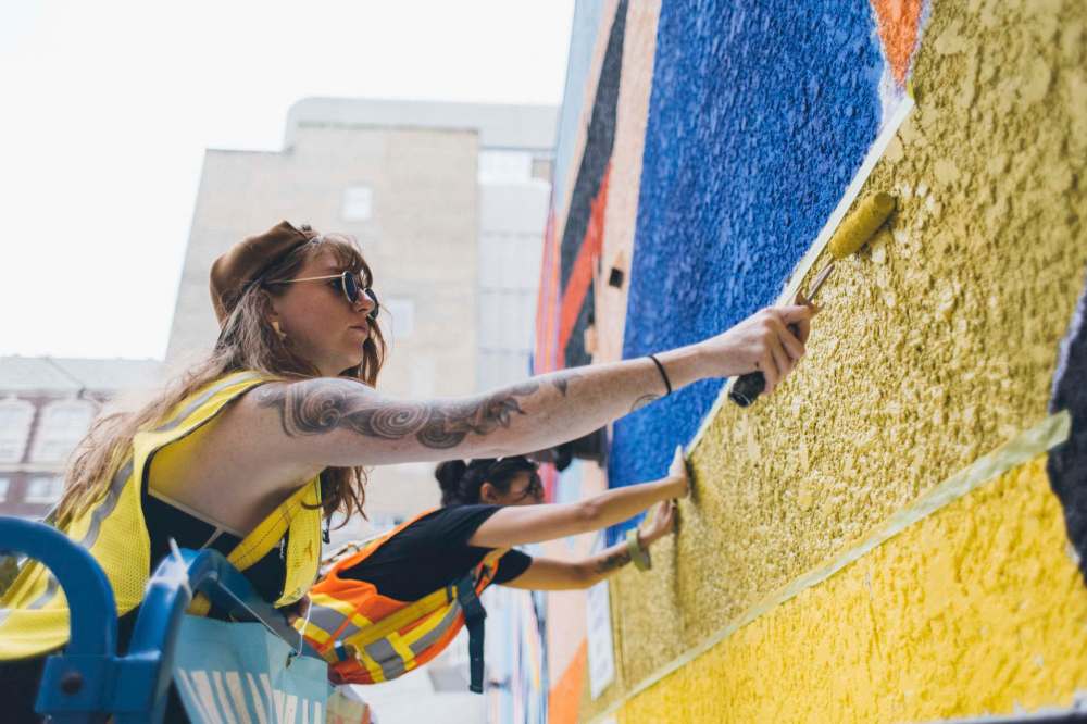 bnb studios Laura Lewis paints Vancouver-based artist Pricilla Yu’s mural Crane Dreamscape behind the Dollarama at 295 Portage Ave.