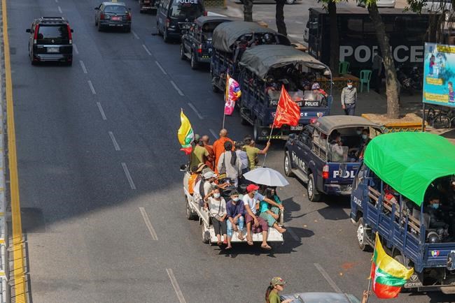 A vehicle with Myanmar and military flags and supporters of the Myanmar military and the military-backed Union Solidarity and Development Party passes by a row of police trucks with police security onboard parked near the Kyauktada police station in Yangon, Myanmar Monday, Feb. 1, 2021. Myanmar military television said Monday that the military was taking control of the country for one year, while reports said many of the country's senior politicians including Aung San Suu Kyi had been detained. (AP Photo)