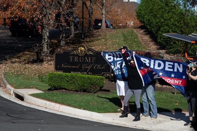 People hold flags supporting Democratic presidential candidate former Vice President Joe Biden as the motorcade carrying President Donald Trump arrives at Trump National Golf Course, Saturday, Nov. 7, 2020, in Sterling, Va. (AP Photo/Evan Vucci)
