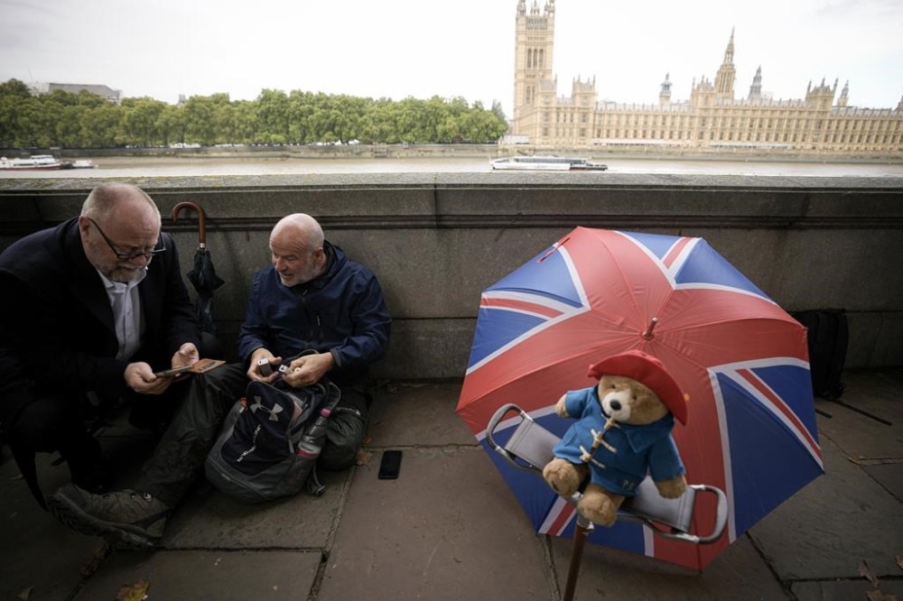 Men sit next to a Paddington bear stuffed toy placed in front of a Union flag umbrella as people wait opposite the Palace of Westminster to be first in line bidding farewell to Queen Elizabeth II in London, Wednesday, Sept. 14, 2022. Queen Elizabeth II, Britain's longest-reigning monarch and a rock of stability across much of a turbulent century, died Thursday Sept. 8, 2022, after 70 years on the throne, at the age of 96. (AP Photo/Andreea Alexandru)