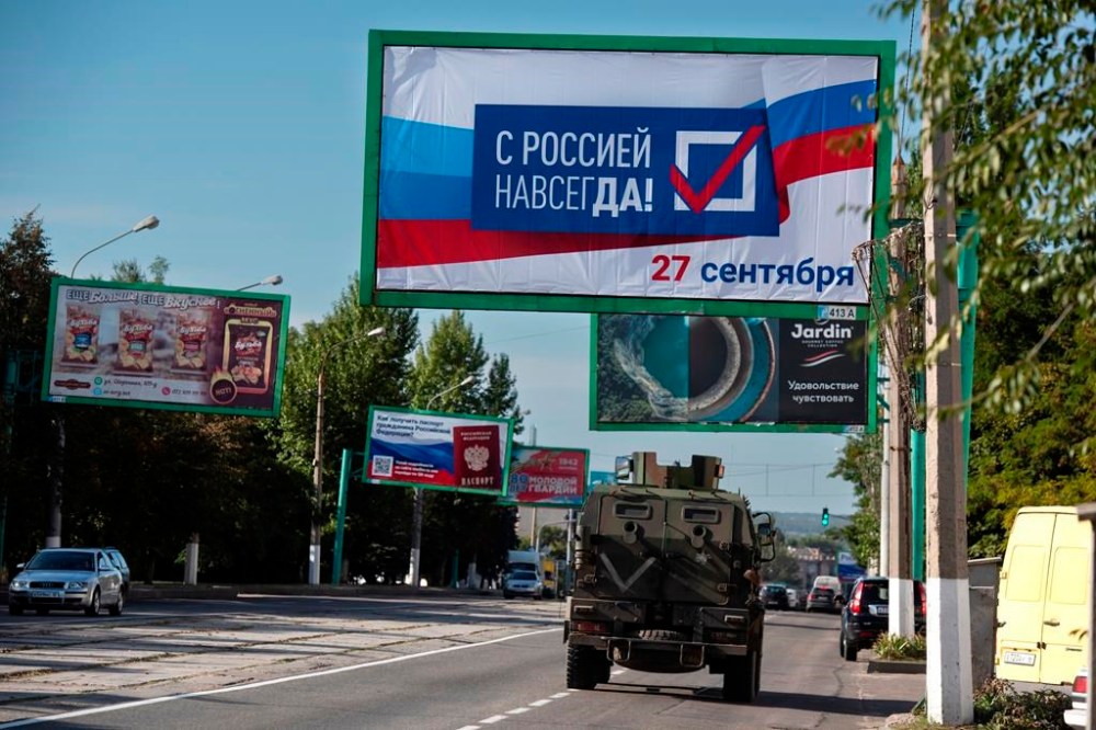 FILE - A military vehicle drives along a street with a billboard that reads:
