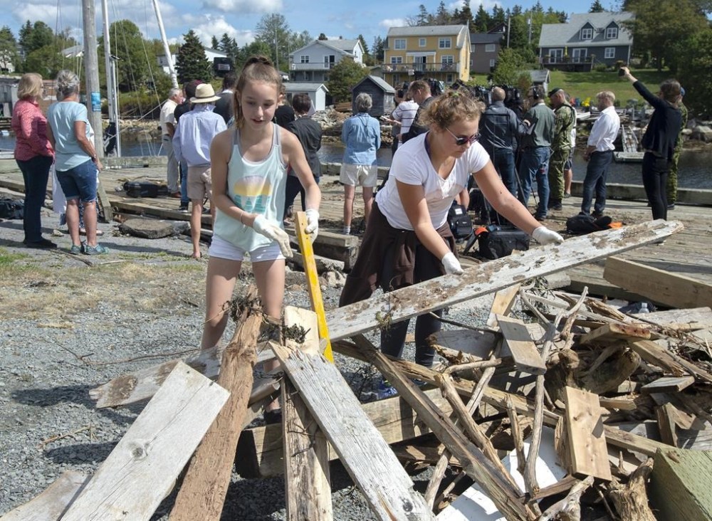 Madelyn Watts, left, and Angelina Taylor clean up debris on the government wharf as Ralph Goodale, minister of Public Safety and Emergency Preparedness and Harjit Sajjan, minister of National Defence, hold a news conference in the background in Herring Cove, N.S. on Tuesday, Sept. 10, 2019. Hurricane Dorian brought wind, rain and heavy seas that knocked out power across the region. THE CANADIAN PRESS/Andrew Vaughan