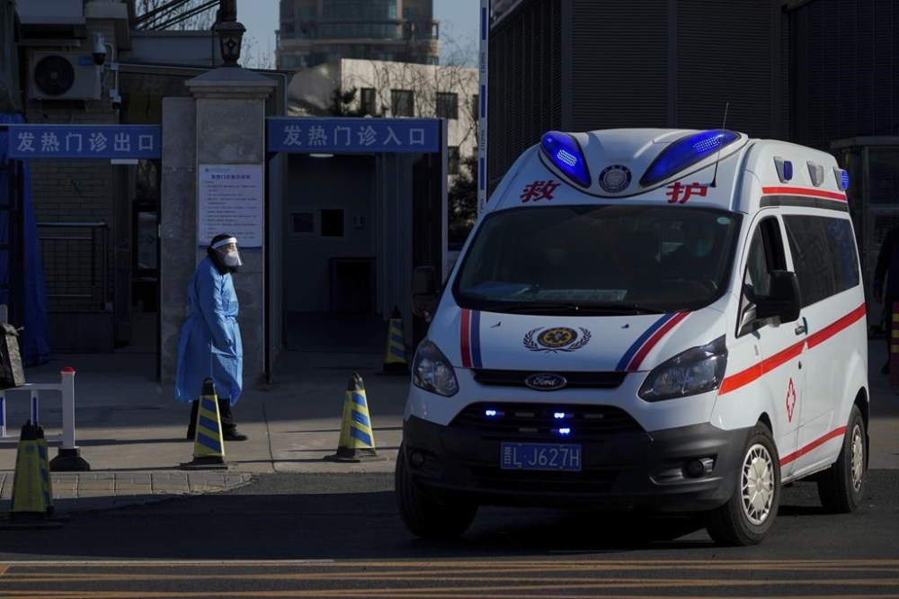 An ambulance passes a worker in protective gear outside a fever clinic in Beijing, Monday, Dec. 19, 2022. Chinese health authorities on Monday announced two additional COVID-19 deaths, both in the capital Beijing, that were the first reported in weeks and come during an expected surge of illnesses after the nation eased its strict